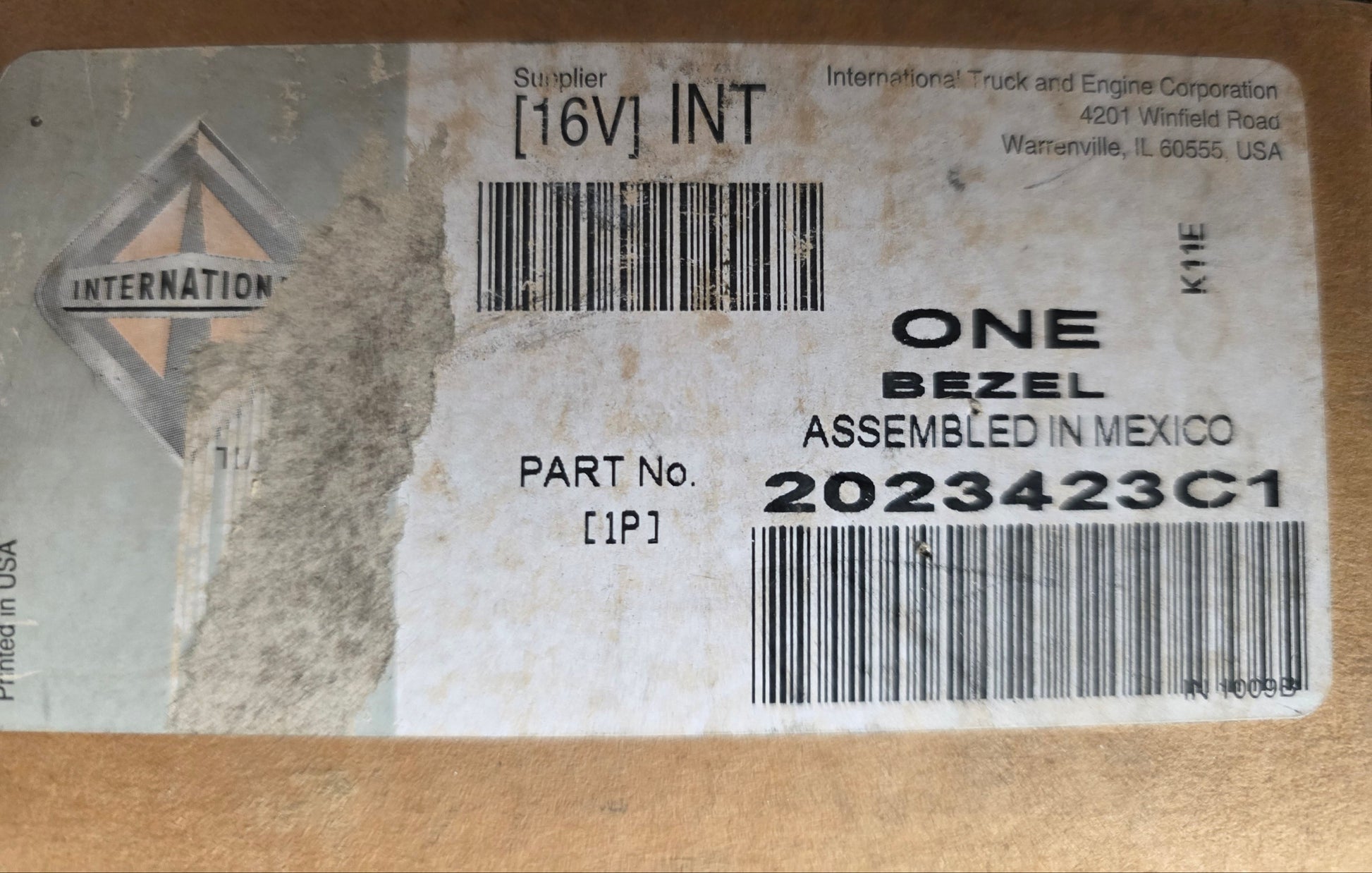 A close-up of a cardboard shipping box with a large white printed label. The label shows the International Brands logo, a barcode, and text including 'ONE', 'BEZEL', 'ASSEMBLED IN MEXICO', and '[16V] INT'.